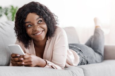 Hayalperest siyah kız kanepede uzanıyor ve akıllı telefonuyla mesajlaşıyor, oturma odasında dinleniyor, çevrimiçi iletişimin tadını çıkarıyor, kapatıyor.