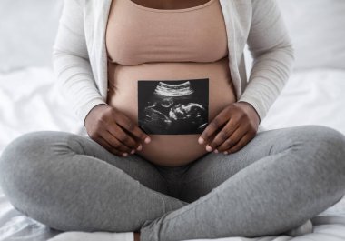 Tanımlanamayan siyahi hamile kadın evde yatağında oturmuş bebeğinin sonografi fotoğrafını gösteriyor. Çocuğunun ilk fotoğraflarını gösteriyor. Doğum gününün keyfini çıkarıyor.