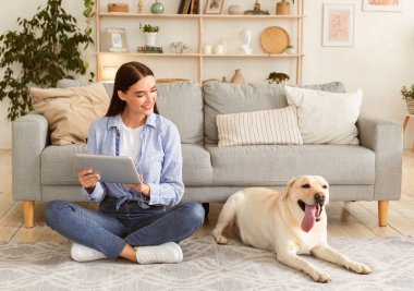 Boş zaman kavramı. Tatmin olmuş kız, elinde tabletle halıda oturuyor ve labradoruna bakıyor.