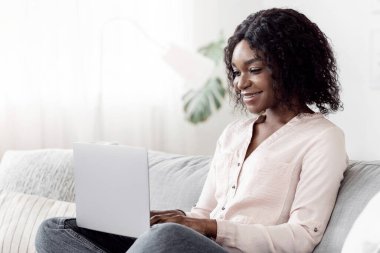 İçişleri Bakanlığı. Gülümseyen Afrikalı kız evde dizüstü bilgisayarda çalışıyor oturma odasında koltukta oturuyor, boş yer var.