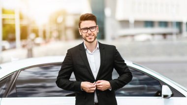 Şehir caddesinde arabasının önünde poz veren kendinden emin genç iş adamının portresi.