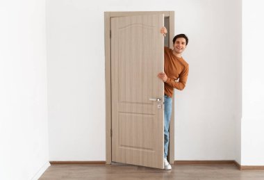 Modern dairenin kapısında duran neşeli genç adamın portresi, milenyum erkek ev sahibi kapıyı tutuyor ve dışarı bakıyor ve gülümsüyor, selamlıyor ziyaretçi, tüm vücut uzunluğu