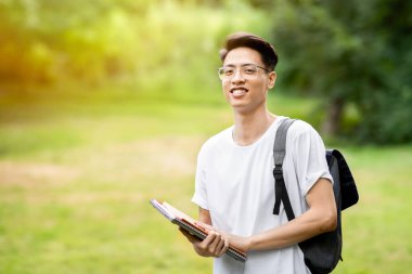 Sırt çantası ve çalışma kitaplarıyla gülümseyen Asyalı öğrencinin portresi üniversite bahçesinde poz veriyor, kameraya bakıyor, mekanı kopyalıyor.
