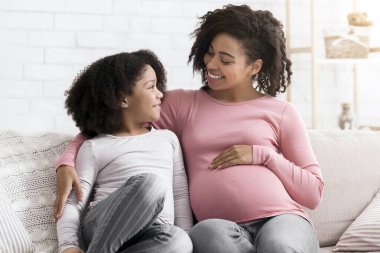 Genç Afrikalı Amerikalı hamile kadın küçük kızıyla evde takılıyor. Kanepede oturuyor ve sarılıyor.