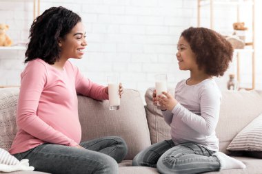 Hamile afro anne ve tatlı küçük kızı birlikte süt içiyorlar, oturma odasında kanepede oturuyorlar, yan gözle bakıyorlar.