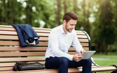 Yakışıklı şirket çalışanı parktaki bankta dinleniyor ve tablet bilgisayardaki interneti tarıyor.