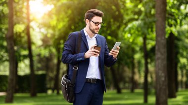 Kendine güvenen genç işadamı cep telefonundan e-postayı kontrol ediyor ve şehir parkında kahve içiyor.