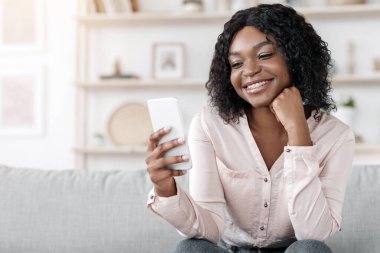 Güzel uygulama. Güzel siyah kadın kanepede oturuyor ve akıllı telefon kullanıyor, evde vakit geçirmekten zevk alıyor, boşluğu kopyalıyor.