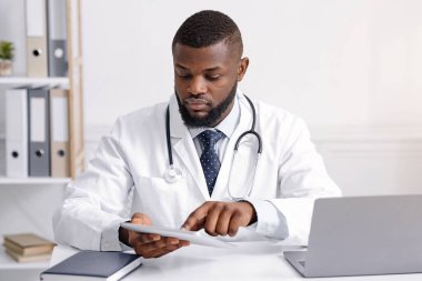 Karantina sırasında ofisinde dijital tablet kullanan konsantre siyah doktor.