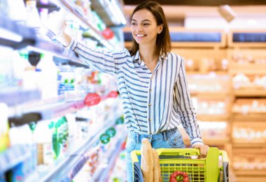 Gülümseyen Neşeli Kadın markette rafların arasında alışveriş arabasıyla geziyor. Marketten alışveriş yapan mutlu bayan, mandıra ürünleri alan buzdolabı reyonunun yanında duran