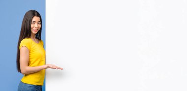 Pozitif Koreli kadın, Beyaz Reklam Kurulu 'nda fotokopi Alanı' nı gösteriyor, boş ilan panosunda avuç içi ile işaret ediyor, mor arka planda duruyor ve kameraya bakıyor.