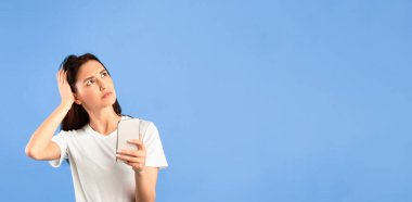 Ne Mesajı? Elinde cep telefonu olan ve kafasını kaşıyarak kafasını kaldırıp mavi stüdyo arka planına bakan düşünceli genç bir kadının portresi.