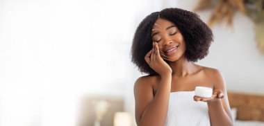 Afro-Amerikalı kadın yüzüne canlandırıcı krem sürerken rahatlıyor, öğleden sonra sakin bir spa ortamında şımartıcı bir cilt bakımı yapıyor. Atmosfer sakin ve huzurlu..