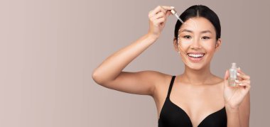 Asyalı kadın yüzüne serumu damlatırken gülümsüyor. Huzurlu bir ortamda gençleştirici bir cilt bakımı uygulaması uyguluyor.