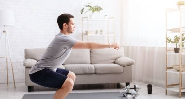 Çalışmak için evde tek başına kal. Oturma odasında çömelme egzersizi yapan genç adam. Evin içinde, boş alanda.