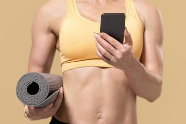 Covid-19 kilitleme ve popüler fitness blogcusu ile online çalışma. Spor giyim bölümündeki zayıf kaslı bayan antrenör minder tutuyor ve akıllı telefona bakıyor, arka planda, panoramada, boş alanda izole edilmiş.