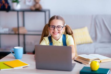 Çalışmaya hazırım. Gözlüklü gülümseyen öğrencinin portresi masada oturuyor, dizüstü bilgisayar kullanıyor ve kameraya bakıyor.