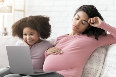 İkinci Hamilelik Sorunları. Evde küçük kızıyla dizüstü bilgisayar kullanırken yorgun düşmüş hamile bir kadının portresi,