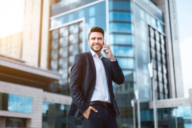 Neşeli genç işadamı ofis binasının önünde mobil telefondan konuşuyor.