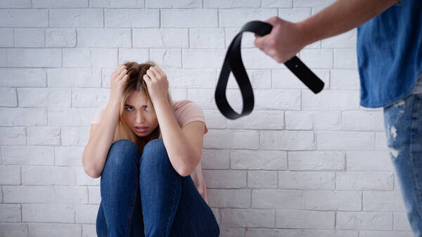 Бедная женщина приседает в страхе перед стеной, в то время как мужчина угрожает ей ремнем, подчеркивая проблему домашнего насилия в отношениях между тысячелетиями.