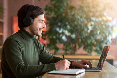 Çevrimiçi yabancı dil öğrenen, kablosuz kulaklık ve dizüstü bilgisayar kullanan, notlar alan, kafenin içini ve boş alanı gören olumlu adamın yan görüntüsü.