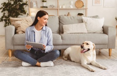 Genç ve güzel bir kadın yerde oturuyor ve dijital tablet kullanıyor. Gülümseyerek köpeğine bakıyor.