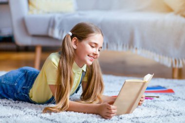 Leisure Concept. Portrait of cute smiling teenage girl lying on the floor carpet at home in living room, holding and reading paper book, enjoying story, copy space. Pupil resting and relaxing
