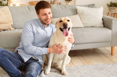 Hayvan Konsepti ile temas. Oturma odasında yerde oturmuş Golden Retriever 'ına sarılan yakışıklı adam portresi. Modern apartmanda, evde vakit geçiren mutlu bir adam.