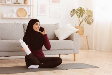Evde antrenman yaptıktan sonra cep telefonuyla konuşan, su içen, oturma odasındaki yoga minderinde oturan mutlu Arap kadın.
