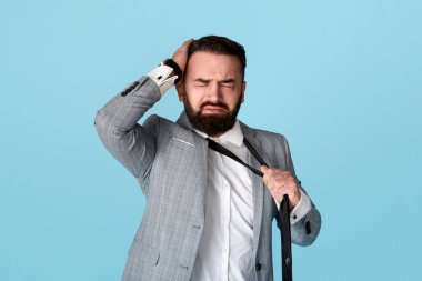 Çaresiz genç ofis çalışanı başını kavrayıp kravatını mavi arka planda çekiyor.