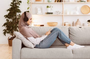 Hafta sonu eğlencesi. Evde akıllı telefon ve kablosuz kulaklıklarla müzik dinleyen tesettürlü rahat bir Arap kızı.