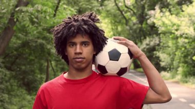 Kıvırcık saçlı, genç bir Afro-Amerikan adam yemyeşil bir parkta futbol antrenmanı yapıyor. Omzunda bir futbol topu tutuyor ve açık havada sağlıklı ve aktif bir yaşam tarzını ön plana çıkarıyor..
