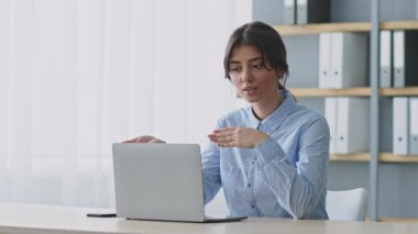 Uzaktan eğitim alan genç bir kadın online bir ders aracılığıyla bilgi paylaşıyor. Modern bir ofis ayarlamış, öğrencilerle laptopuyla iletişim kuruyor..