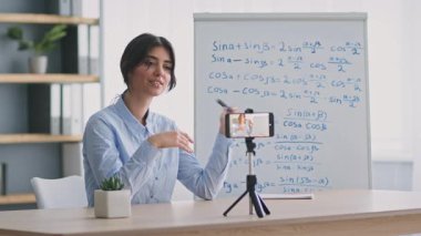 Genç bir kadın online bir ders veriyor, telefonuyla öğrencilerle iletişim kuruyor. Matematik kavramları, o seyircilerle coşkuyla uğraşırken arkasındaki tahtada resmedilmiştir..