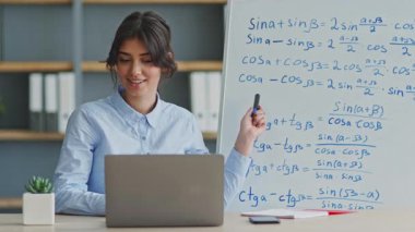 Genç bir öğretmen evde dizüstü bilgisayar kullanarak uzaktan matematik dersi veriyor. Denklemlerle dolu bir tahtadaki kavramları açıklarken öğrencilerle takılıyor..