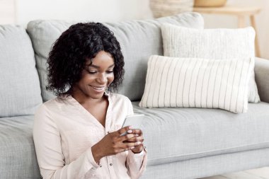 Neşeli siyah kadın evde akıllı telefon ve kahveyle rahatlıyor, arkadaşlarıyla mesajlaşıyor ya da sosyal ağlara takılıyor, oturma odasında oturuyor, izin gününün keyfini çıkarıyor ve ev eğlencesi yapıyor.