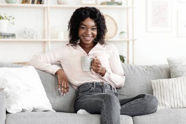 Hafta sonu tatili. Pozitif siyah kadın evde çay içiyor ve koltukta dinleniyor sıcak içecekle bardağı tutuyor ve kameraya gülümsüyor.
