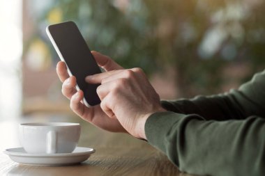 Kafede akıllı telefon kullanan, kahve içen, yan gören bir adam.