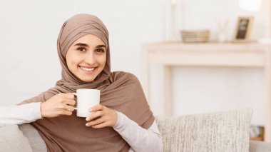 Yurtiçi rahatlama. tesettürlü gülümseyen Arap kız bir fincan kahveyle rahatlıyor, ev konforunun tadını çıkarıyor, kamera karşısında gülümsüyor, panorama ve fotokopi alanı.
