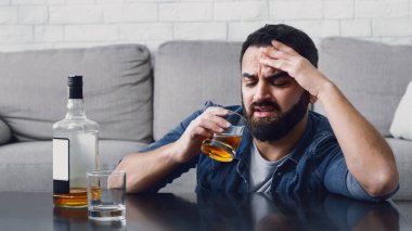 Alkol bağımlılığı sorunu. Sakallı üzgün adam bir bardak viskiye bakar, masanın yanında bir şişe ve oturma odasının içinde cam vardır.