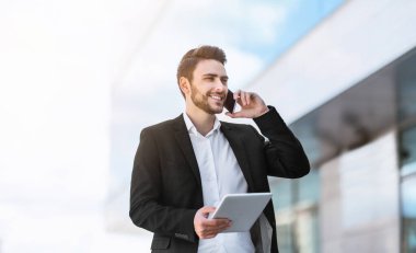 Meşgul genç girişimci cep telefonuyla iletişim kuruyor ve şehirdeki tablet bilgisayarda panorama