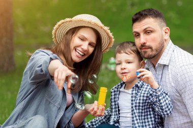 Piknik ailesi eğlencesi. Yazın kırsal bölgede anne ve babasıyla sabun köpüğü üfleyen sevimli çocuk, panorama.