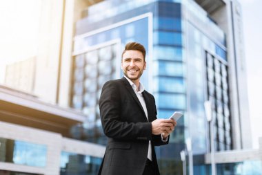 Modern metropolün şehir merkezinde akıllı telefonlu gülümseyen bir CEO.