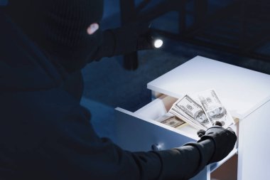 Maskeli soyguncu çekmeceden para çalıyor, el feneri kullanıyor, omzunun üzerinden yakın plan portre çekiyor, arkadan bakıyor.