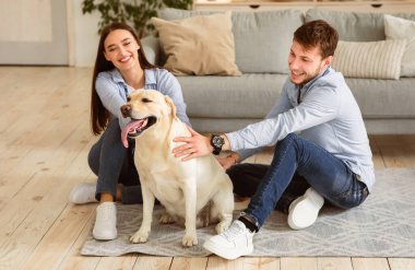 Mutlu Aile Kavramı. Modern bir dairede yerde oturup köpekle vakit geçiren neşeli eşlerin portresi. Erkek ve kadın sahipleri evde labrador hayvanıyla oynuyorlar.