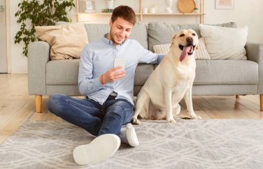 Yaşam tarzı kavramı. Akıllı telefonu kullanan gülümseyen bir adamın portresi halının üzerinde Labrador 'uyla oturmuş, köpeğini okşuyor. Adam evde vakit geçiriyor, sosyal medyada sohbet ediyor, mesaj yazıyor ya da mesaj okuyor.