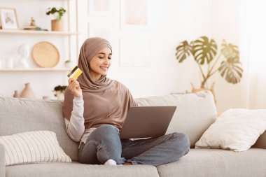 İnternetten alışveriş. Laptop ve kredi kartıyla tesettürlü neşeli Arap kız internetten mal alıyor, evdeki koltukta oturuyor, yer kopyalıyor.