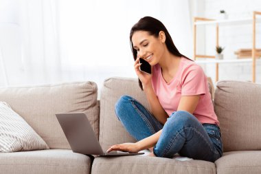 Evde dizüstü bilgisayar ve cep telefonuyla çalışan serbest çalışan kadın portresi. Mutlu kadın internette sörf yapıyor, akıllı telefondan konuşuyor, kanepede oturuyor, oturma odasında dinleniyor, mekanı kopyalıyor.
