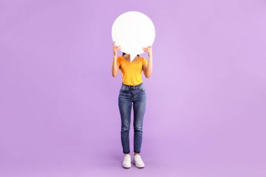 Tanımlanamayan bir kadın kafasının önünde boş bir konuşma balonu tutuyor. Mor stüdyo duvarı, tam boy, dikey çekim.