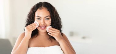 Kıvırcık saçlı bir kadın cilt bakımını yaparken gülümsüyor, yüzüne pamuk pedler takıyor. Yumuşak ışıklandırma, onun rahatlatıcı ifadesini vurguluyor, özbakımını ve tazelemesini vurguluyor..